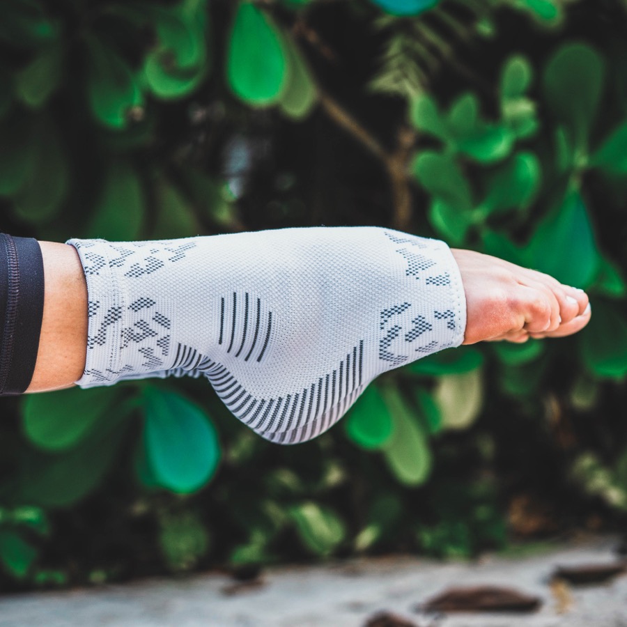 Close-up of a white NanoSocks compression sock on an outstretched foot in daylight against green foliage, showing the knit texture, the compression chevrons over the arch and heel, and the open end where the bare toes come out.
